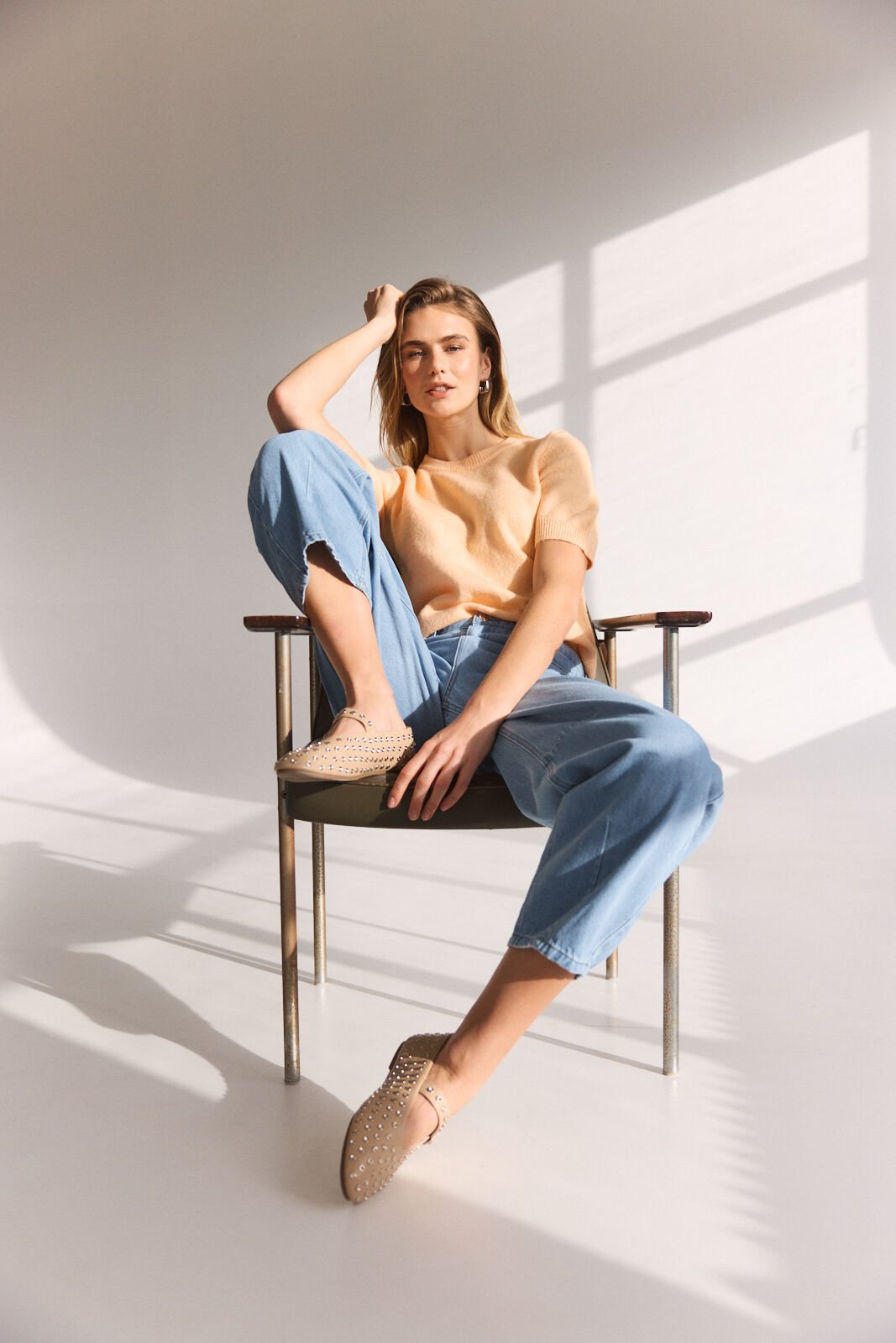 Model sitting in a chair in the sunlight wearing a pastel orange pullover and a light jeans